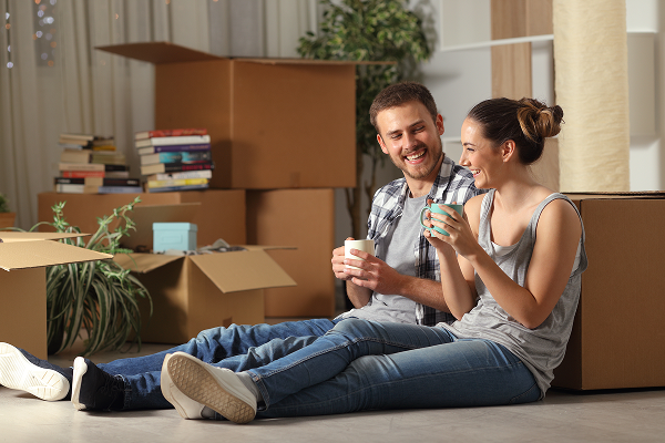 Smiling couple sitting on the floor reviewing home renovation blueprints with paint swatches and a coffee mug, surrounded by paint cans and a ladder.