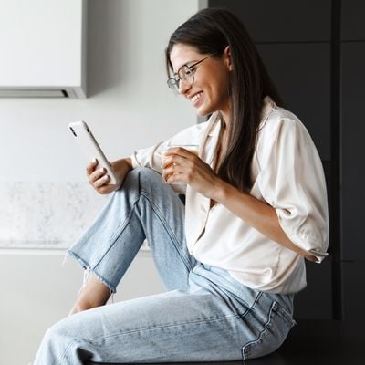 Young woman sitting on her kitchen counter, looking at her phone while  drinking juice.