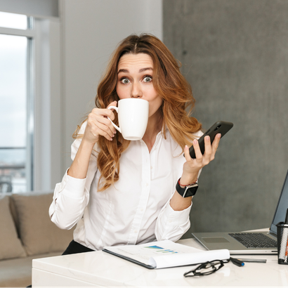 Woman in her office, drinking coffee and looking at her mobile phone.