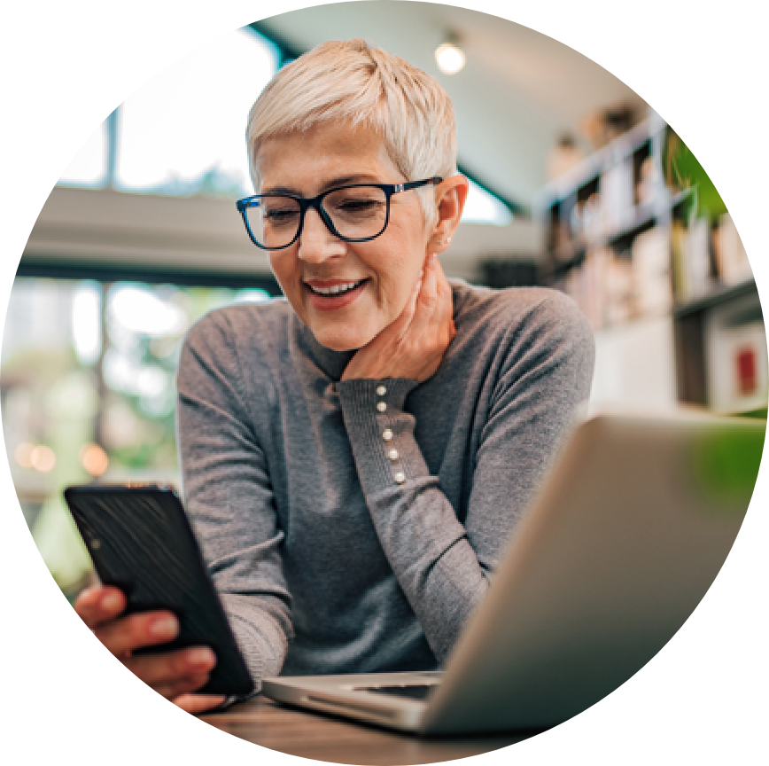 A sharply dressed middle-aged woman sits at a laptop and looks at her smartphone.