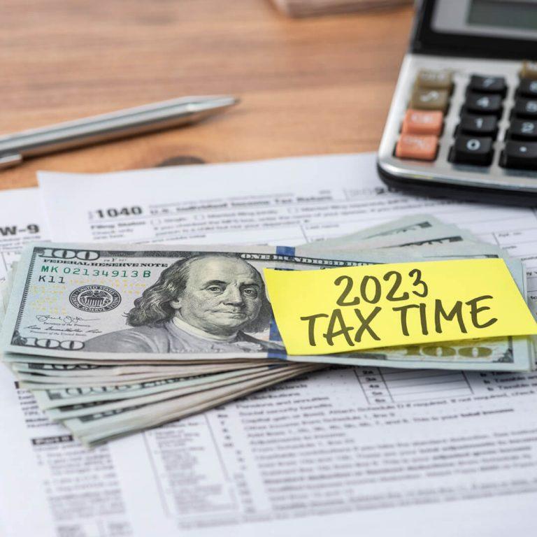 Tax forms and calculator on desk, with hundred dollar bill and "2023 tax time" post it note.