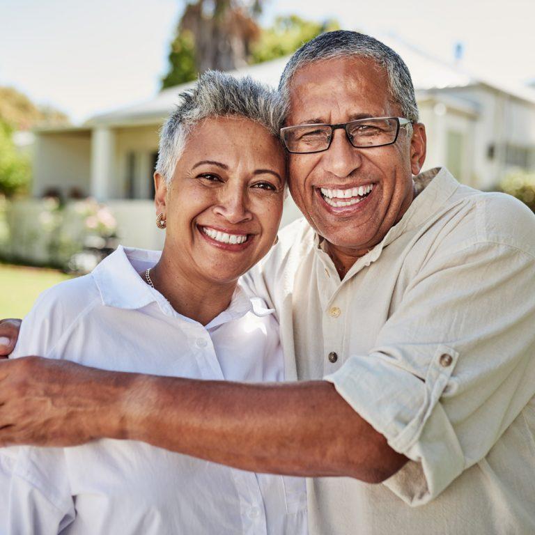 Older smiling couple standing outside a 55+ community home.