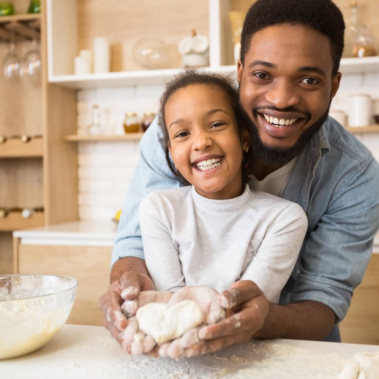 Father and daughter in kitchen baking, looking at the camera and smiling.
