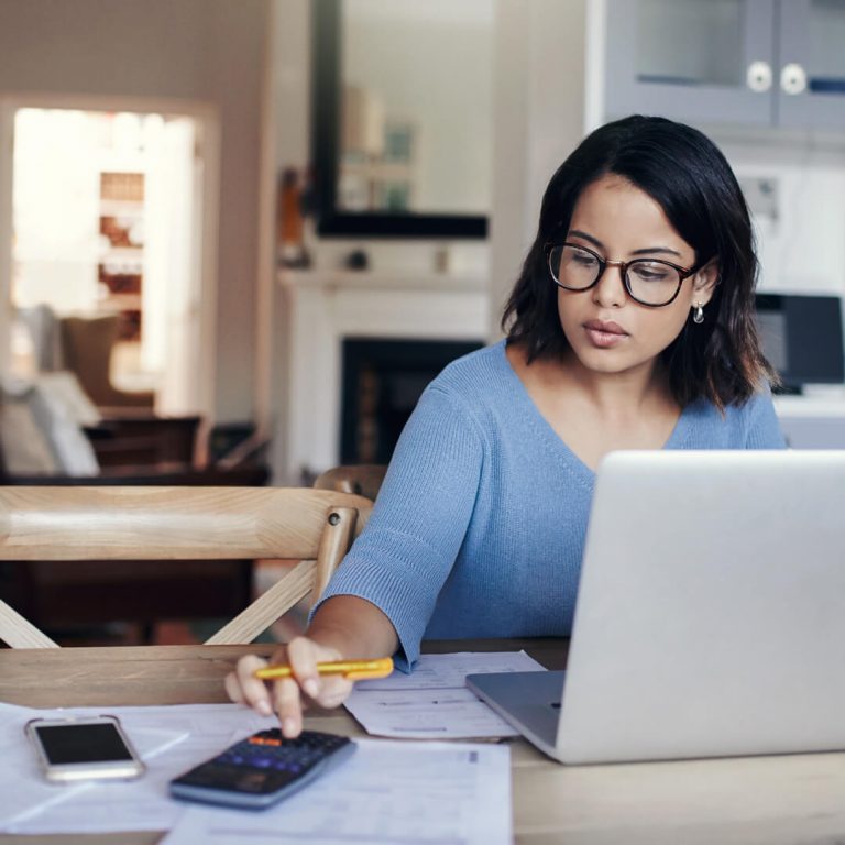 Woman sits at her desk with laptop and calculator, working on her budget.