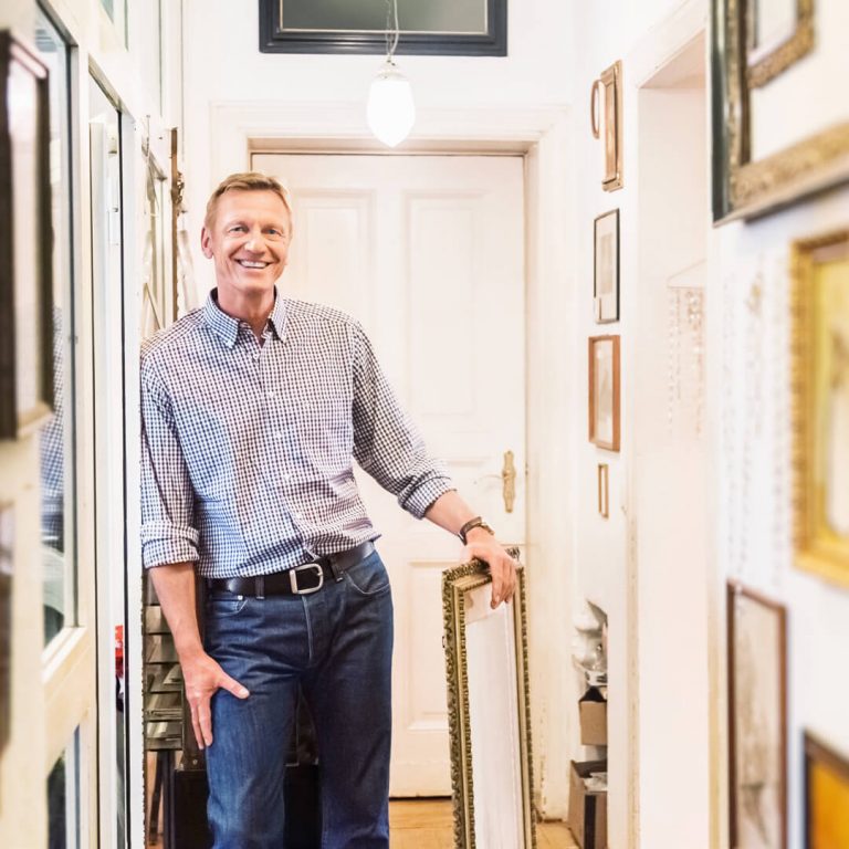 Man smiling in hallway with his private art collection framed and displayed on wall.