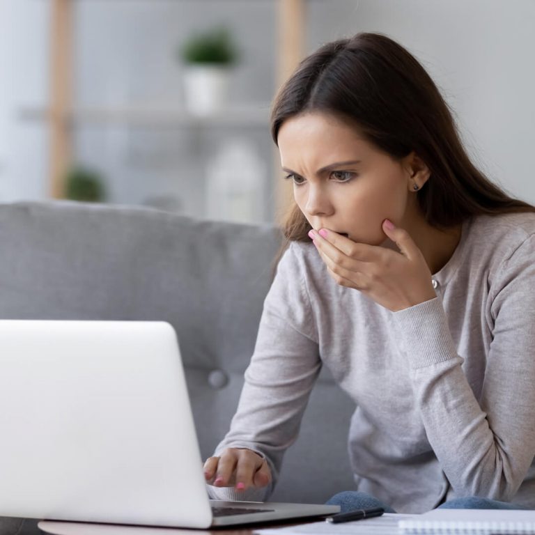 Young woman looks at computer screen in horror, victim of college degree scam.