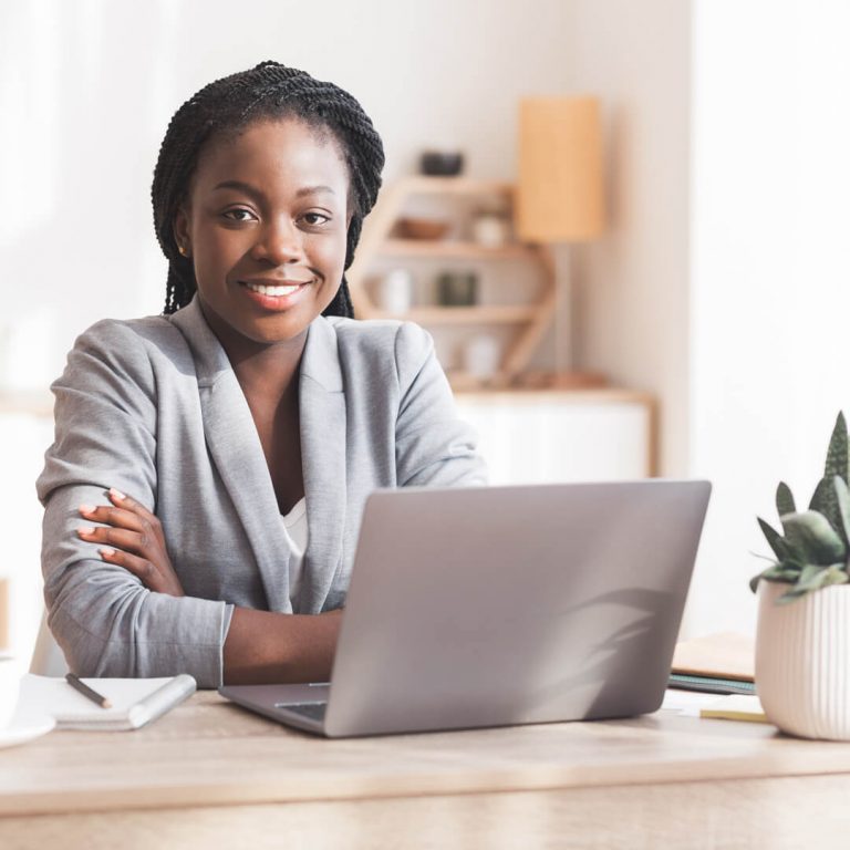Businesswoman seated behind desk, looking out from behind her laptop, smiling confidently at camera.
