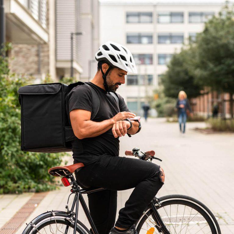 Gig worker (delivery man on bicycle) checks food order on his wearable, with food strapped to his back.