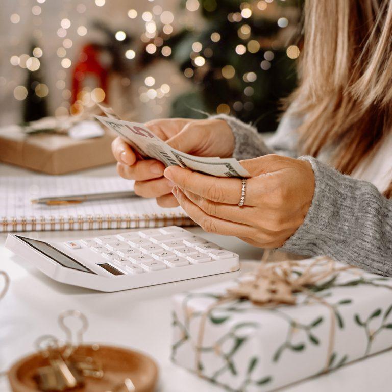 Woman budgeting money for the holidays, holding cash, looking at calculator and near gifts.