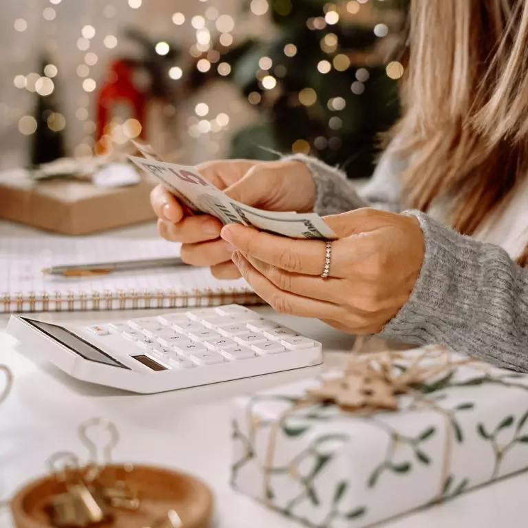 Woman budgeting money for the holidays, holding cash, looking at calculator and near gifts.