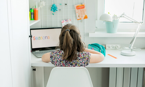 Young girl sitting at desk at home classroom, with laptop and school supplies.