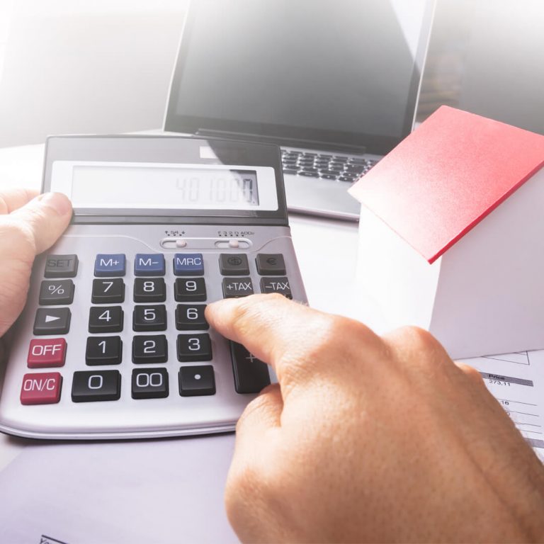 Closeup of hands punching numbers into calculator, wooden house to side.