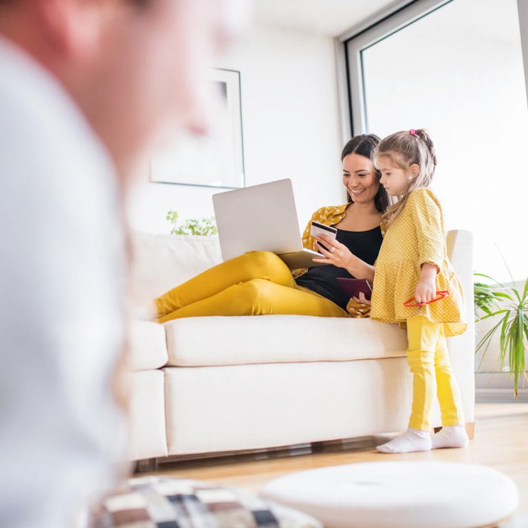 Mother and child in living room; daughter watches her mother purchase something online with a credit card.