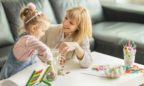 Mother and young daughter seated in living room, putting change into a jar.