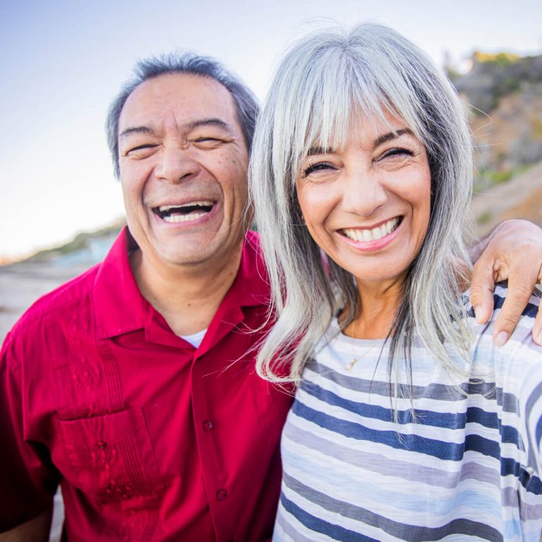 Retired couple smiling at the camera standing on beach.