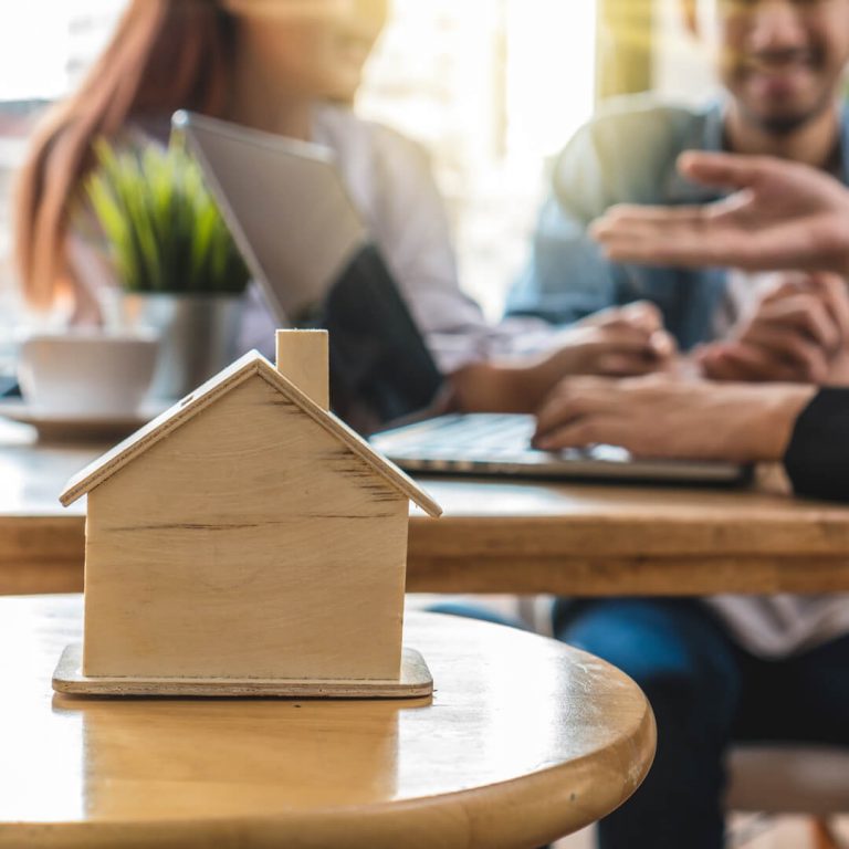 Couple in mortgage broker's office, wooden house on desk in foreground.