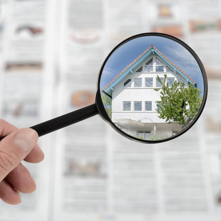Hand holding magnifying glass over real estate ads, honing in on large white house.