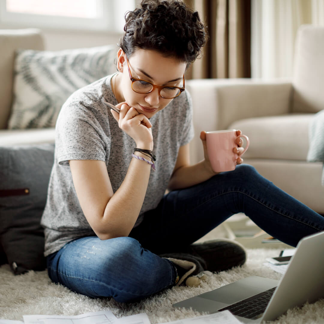 Young woman seated on floor in living room with laptop, reviewing her finances and student loans.