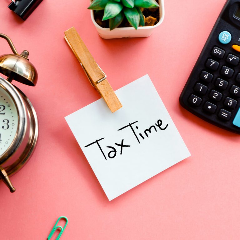 Post it note reading "Tax Time" on pink table with clock and calculator.