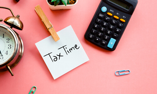 Post it note reading "Tax Time" on pink table with clock and calculator.