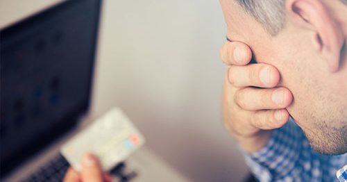 Upset man with face in one hand, credit card in another, worried he may be the victim of identity theft.