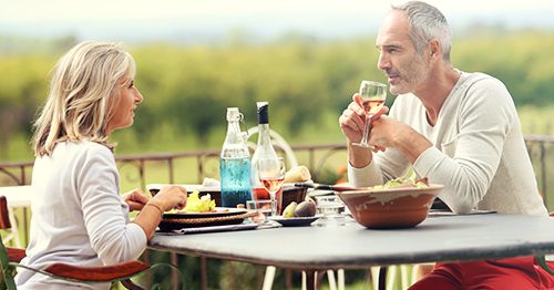 Recent retiree couple sit outside enjoying lunch and wine overlooking a vineyard.