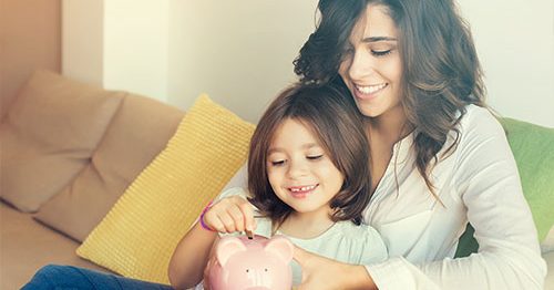 Mother and daughter sitting on couch smiling and adding coins to a piggy bank.