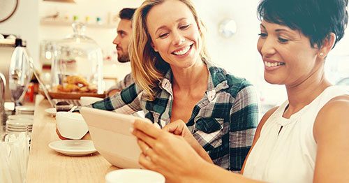 Two 40-year-old women smiling at counter enjoying coffees and looking at laptop.