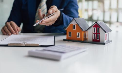 Loan Subordination Agreement on table, next to a model home, and a man motioning camera to sign agreement.