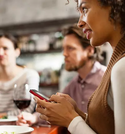 A young woman uses her Quorum mobile banking app on her cell phone at lunch with friends.
